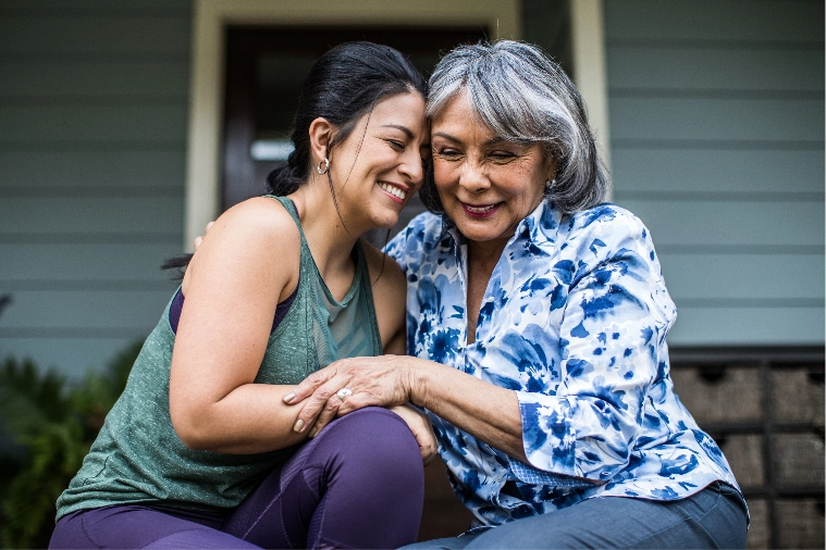 Mother and daughter smile as they embrace while sitting in front of a house.
