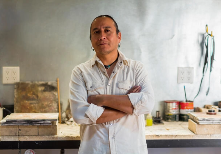 A man, whose face shows confidence, crosses his arms as he leans on his workbench.